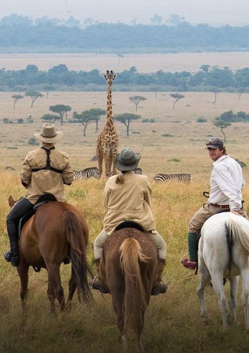 Three horseback riders observe a giraffe on an African savanna safari with distant mountains