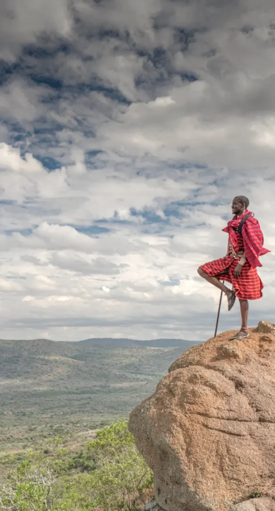 Maasai warrior in traditional red shuka standing on rock outcrop overlooking East African savanna landscape