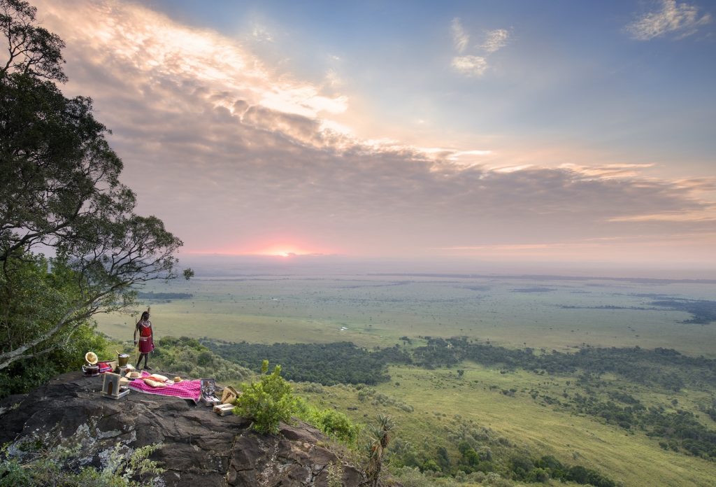 Luxury safari guests enjoying sundowner drinks on rocky outcrop overlooking Masai Mara savanna at sunset